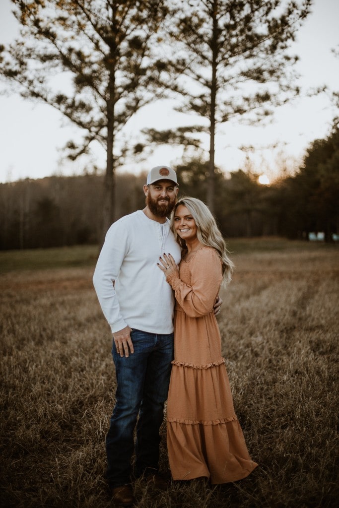 Michael and Madison Wood smiling and embracing in a grassy field at sunset with tall pine trees in the background, featured for Dameron Plumbing & Heating Inc.